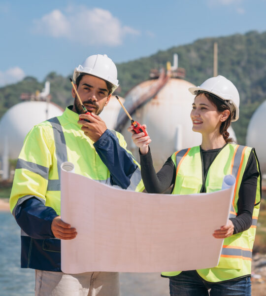 Two engineers in safety gear with blueprints discussing work in front of industrial storage tanks on a clear day.